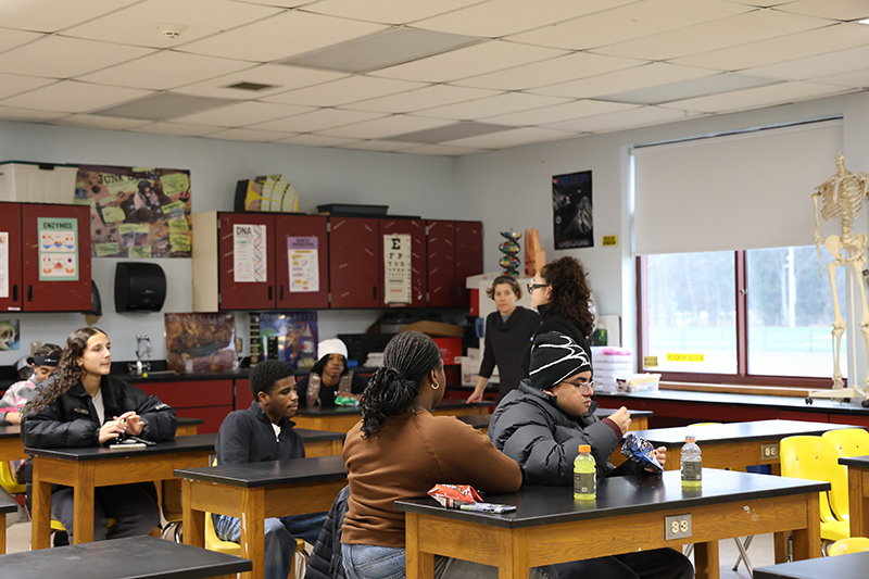  woman stands amongst high school students who are sitting at desks as they talk.