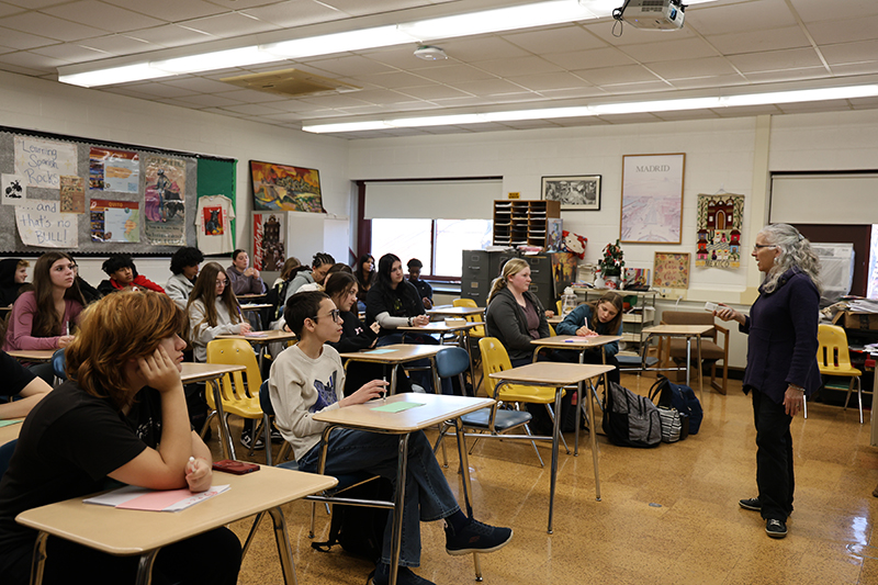 A woman stands at the front of a classroom and speaks to high school students, who are sitting at desks.