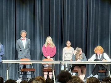 Five high school kids stand and sit on a stage.