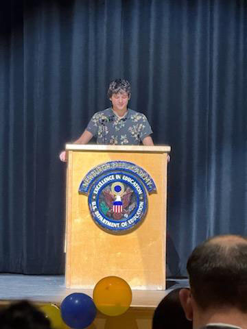 A high school student stands at a podium speaking to a group.