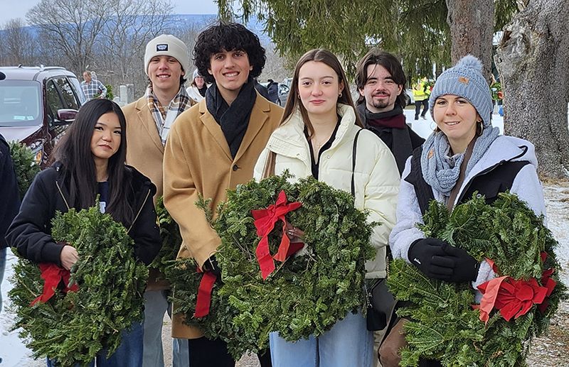 Six high school students dressed for winter weather stand together smiling and holding evergreen wreaths.