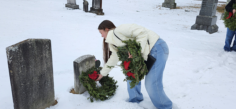 A high school student lays an evergreen wreath at a grave.
