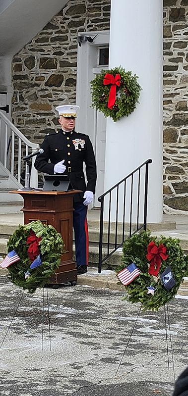 A service member dressed in his dress uniform speaks behind a podium. There are a few wreaths near him. 