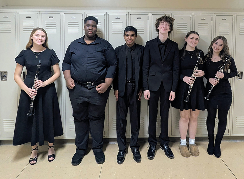A group of six high school students, all dressed in black and some holding musical instruments, stand together and smile.