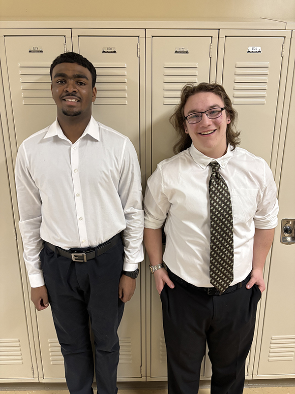 Two high school boys stand together, wearing black pants and white dress shirts. They are smiling.