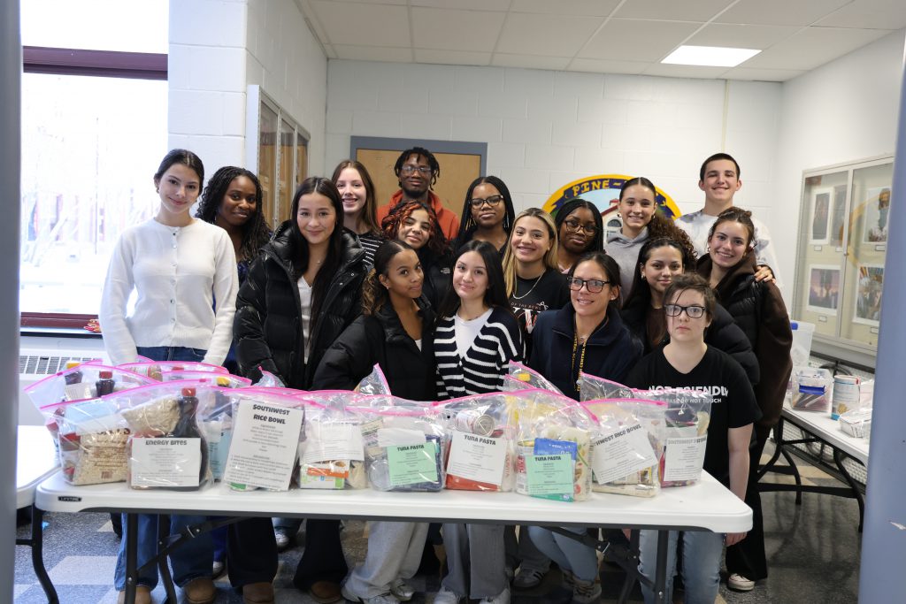 A group of high school students stand behind a table that is filled with meal kits they put together. They are smiling.