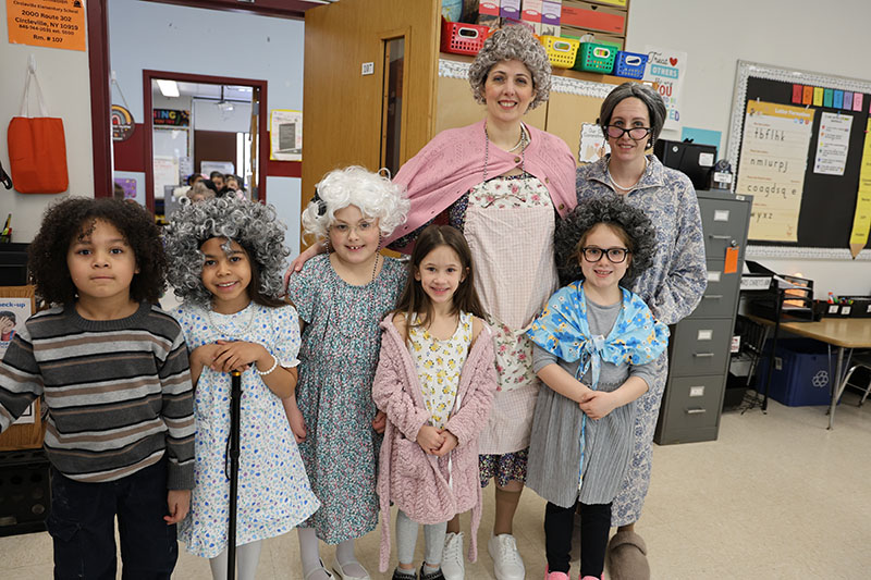 Two women dressed with gray wigs and older lady clothes stand with five first-grade children who are also dressed as old people.