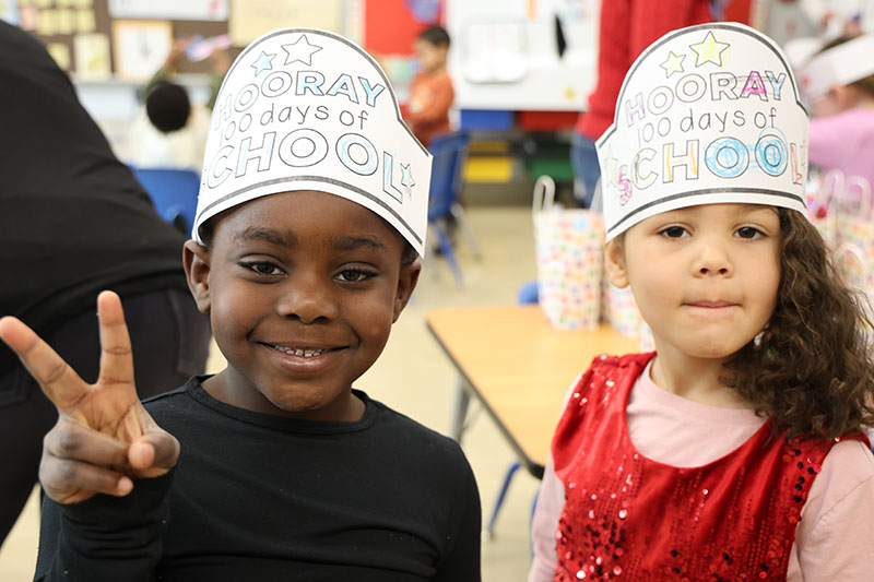 Two younger kids smile as they wear paper hats they made that say Hooray 100 days of school.