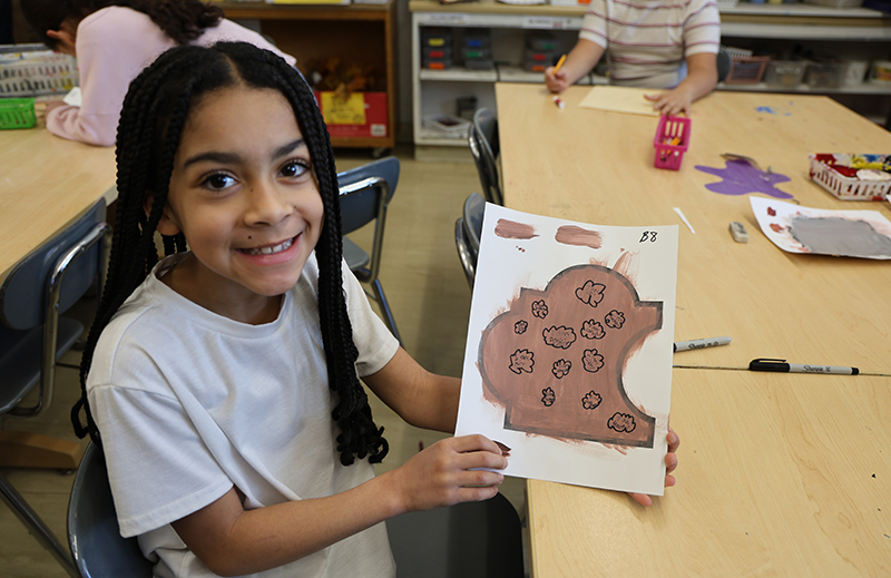 A fourth-grade girl with long dark hair smiles and holds up a puzzle piece she made.