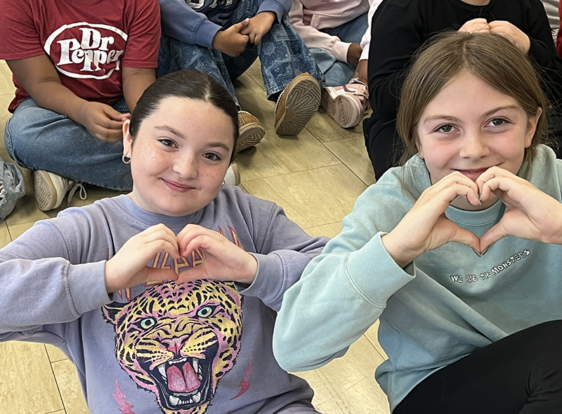 Two girls smile and make the shape of hearts with their hands.