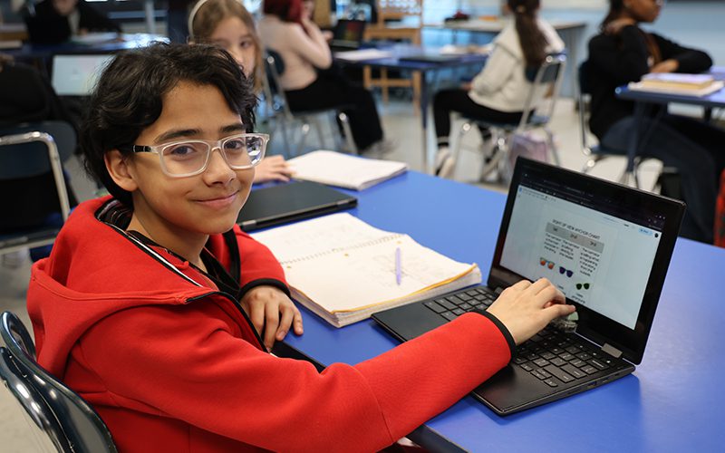 A sixth-grade student with dark hair and glasses, wearing a red hoodie, sits at a desk with a Chromebook. He is smiling.