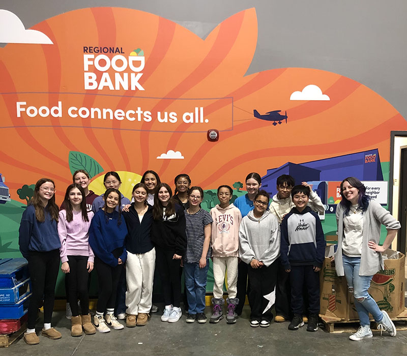 A group of 15 middle school kids and a teacher on the right stand together in front of a large backdrop that says Regional Food Bank. Food connects us all.