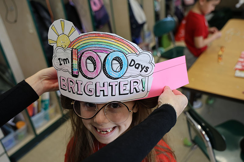 A younger elementary students smiles as she gets her paper hat on that says I'm 100 days brighter!