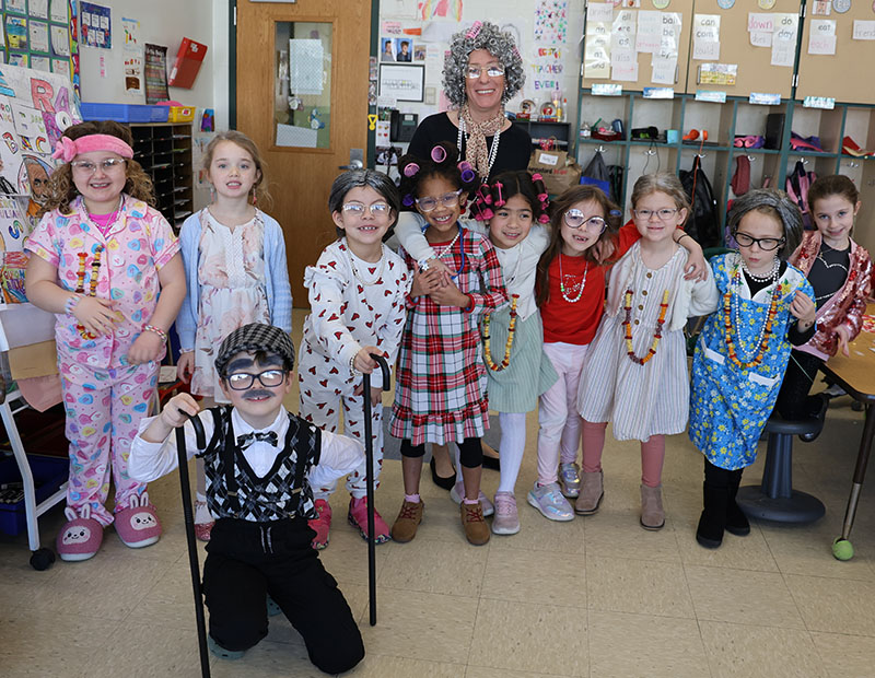 A woman and several younger elementary students stand together all dressed as old people.