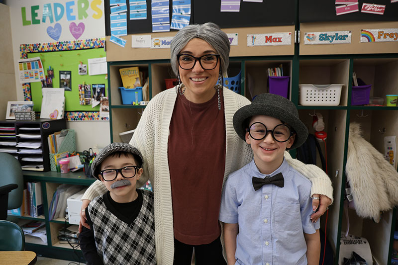 A woman dressed as an old woman stands and smiles with two elementary age boys who are also dressed as old men.