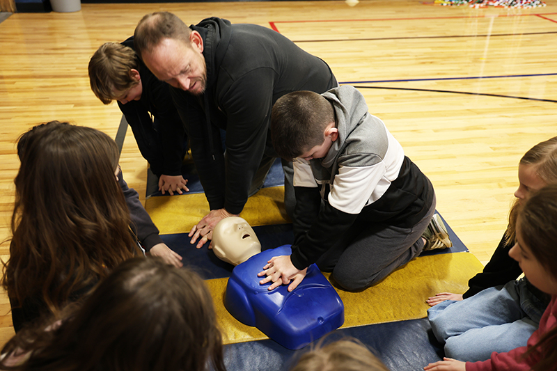 A man leans in as a fourth-grade boy performs CPR on a manikin.