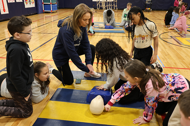 A woman leans in to show fourth-graders the right way to perform CPR.
