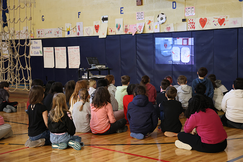 A group of fourth-grade students sit on a gym floor watch a video about hearth health.