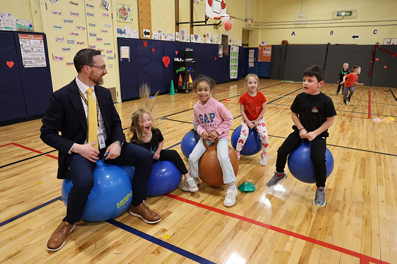 A man in a suite and tie sits on a hippity hop ball while four kindergarten kids hop on them.