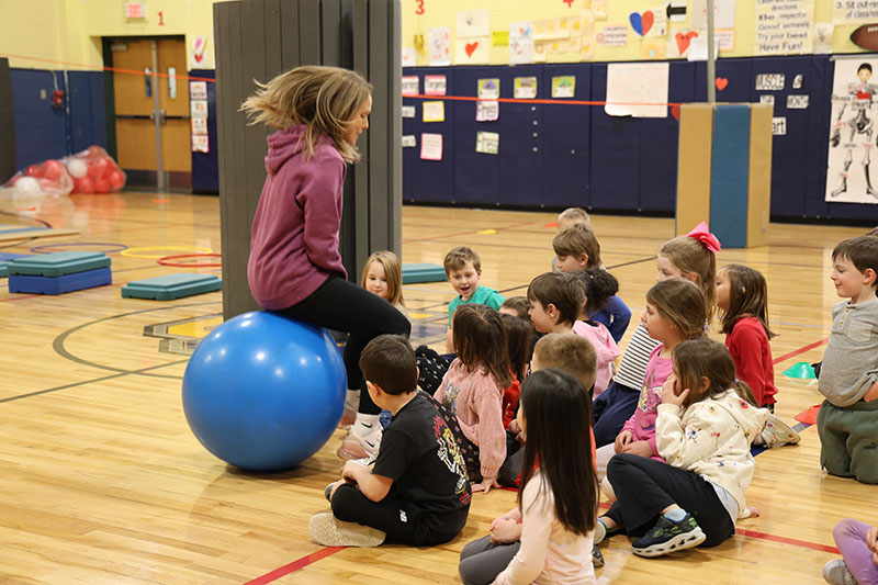 A woman sits on a blue hippity hop ball and bounces, showing a group of kindergarten kids how its done.