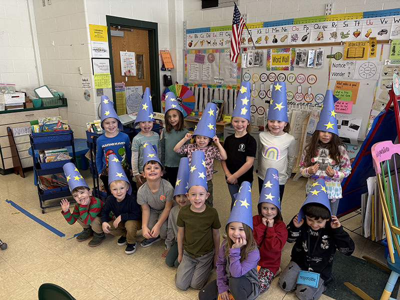 A group of kindergarten kids, about 20, with blue paper wizard hats on. The hats have stars on them.