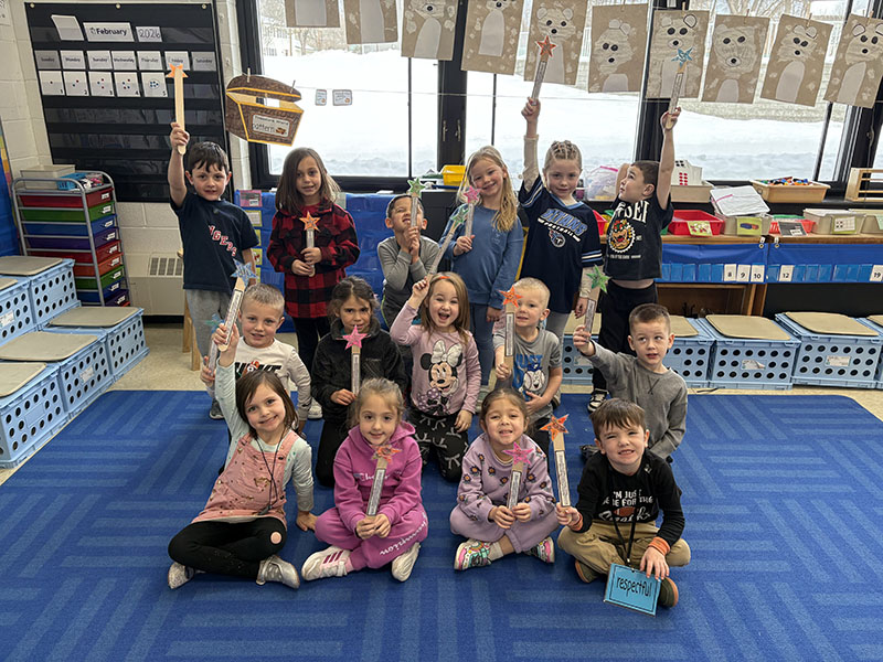 A group of 15 kindergarten kids sit or stand in three rows. Each is holding a magic wand they made.