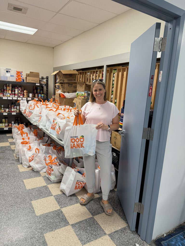 A woman smiles at the door of a food pantry. She is holding a bag of food. Written on the bag is Regional Food Bank.