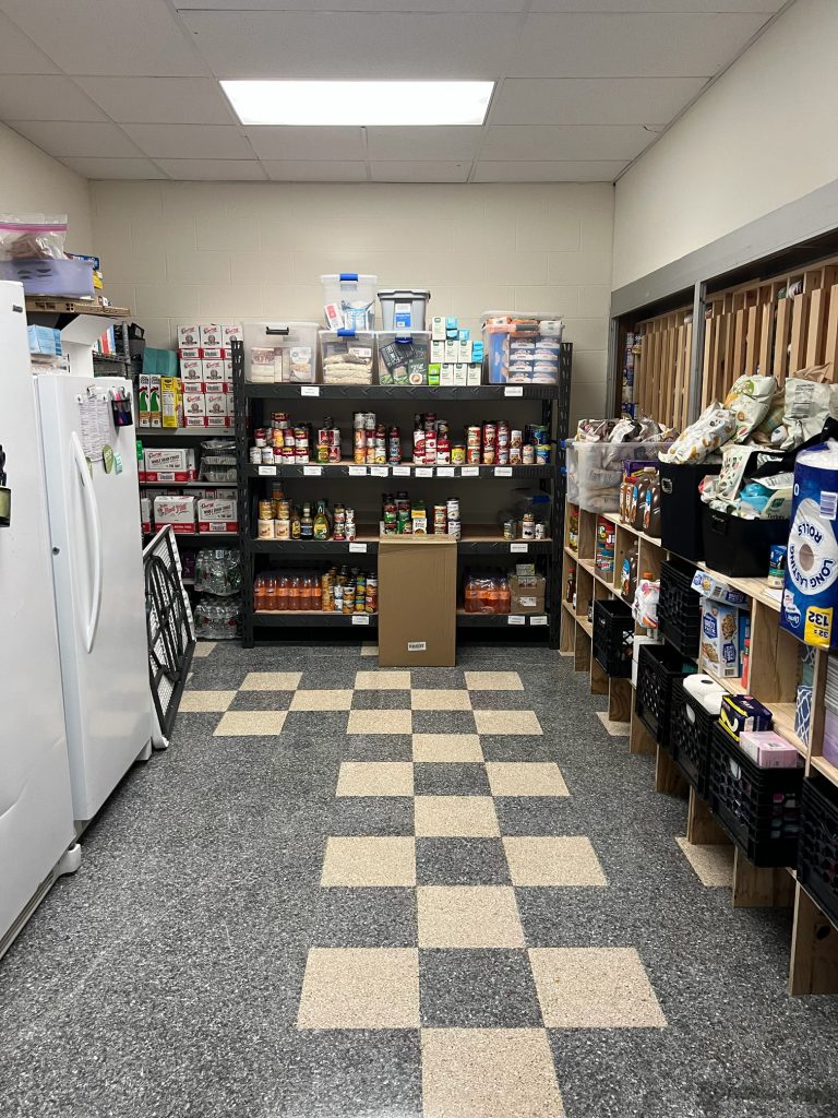 Looking in the door of a food pantry. there are refrigerators on the left, shelves straight ahead and shelves on the right.