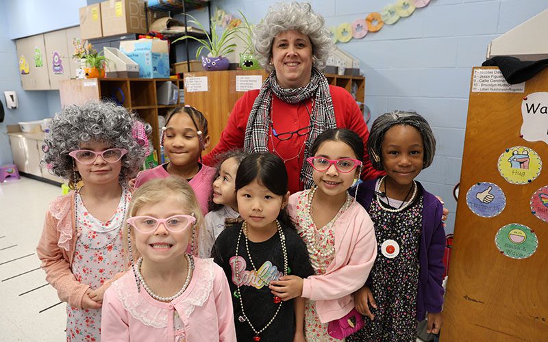 PAK 100th day all the oldies web A woman dressed as a very old woman stands with seven first-graders who are also dressed as 100-year-old people.
