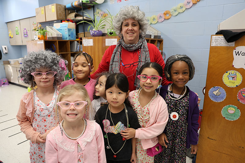 A woman dressed as a very old woman stands with seven first-graders who are also dressed as 100-year-old people.
