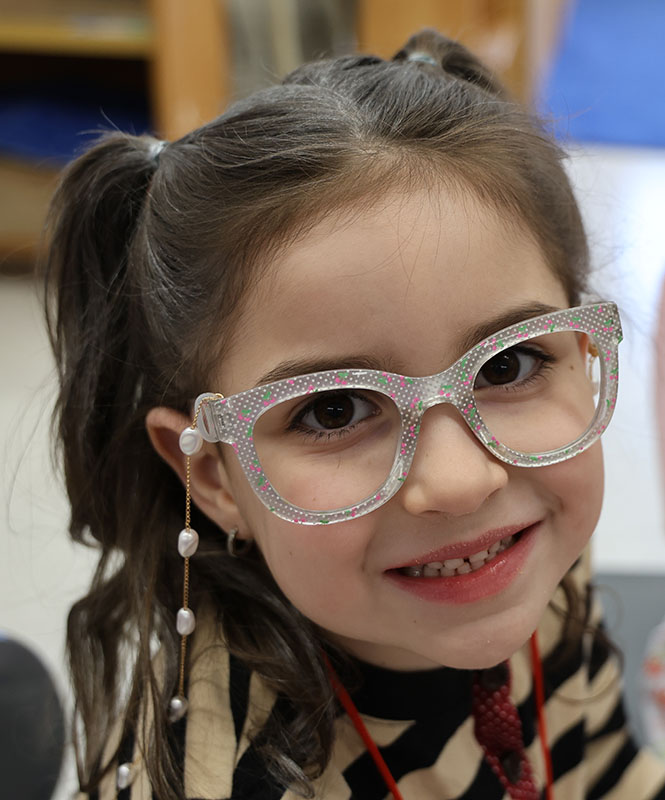 A young elementary girl is dressed like an old lady with glasses and a pearl chain to hold them around her neck.