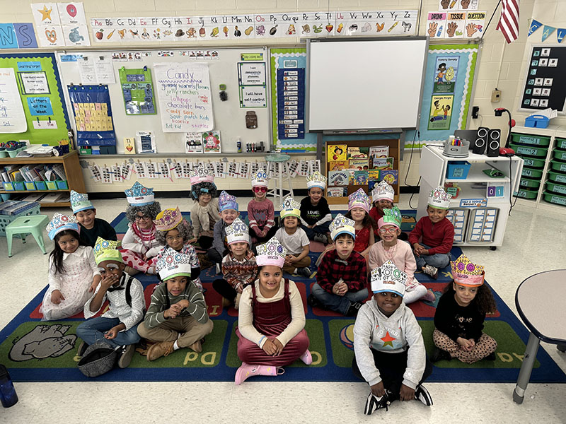 A group of young elementary students sit with hats that say Happy 100th day of school.