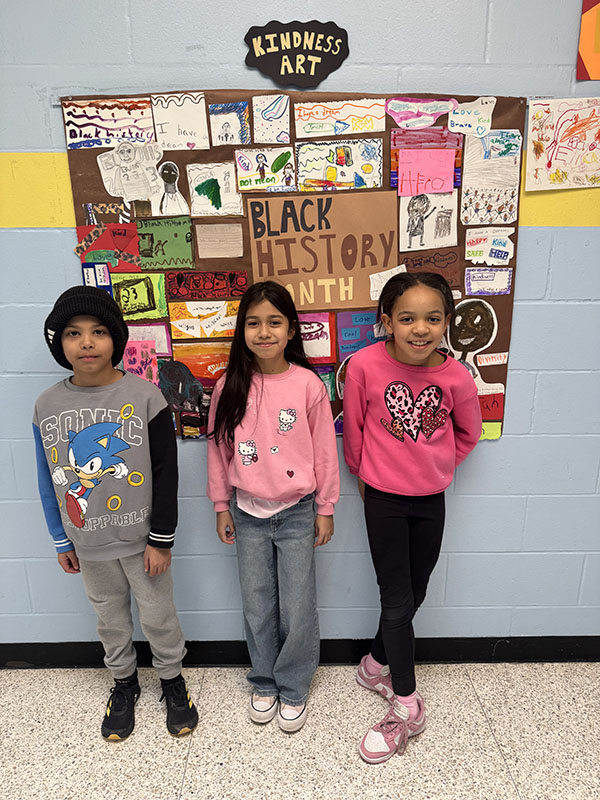 Three elementary kids stand in front of a board filled with art. It says Black History Month.