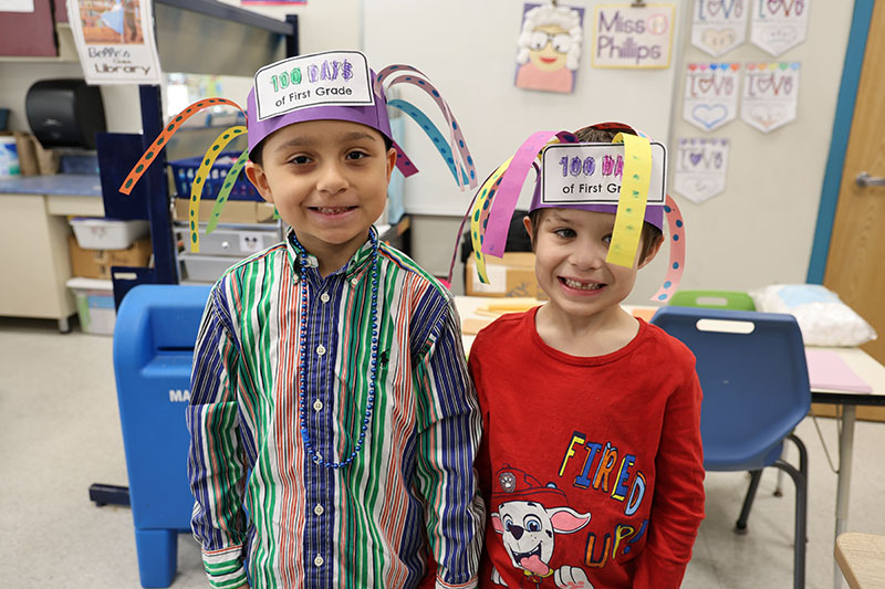 Two younger elementary boys smile while wearing silly hats that have strips of paper in different colors hanging over their heads. Each strip of paper has 10 dots on it.