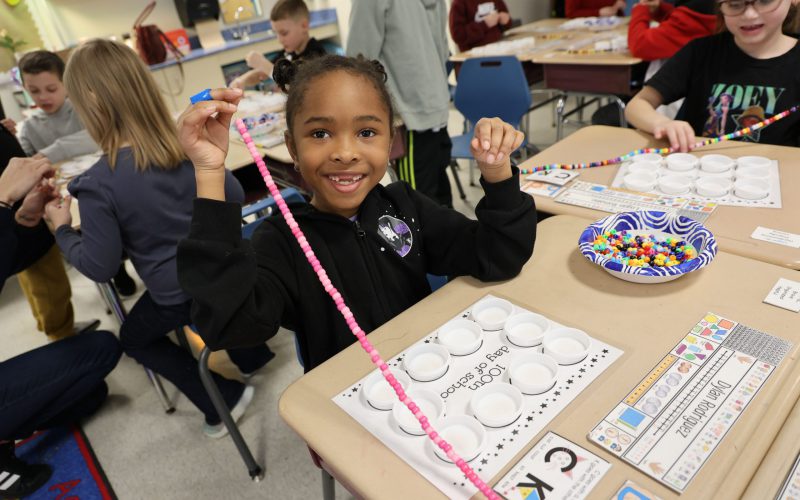 A young elementary girl smiles as she strings 100 pink beads.