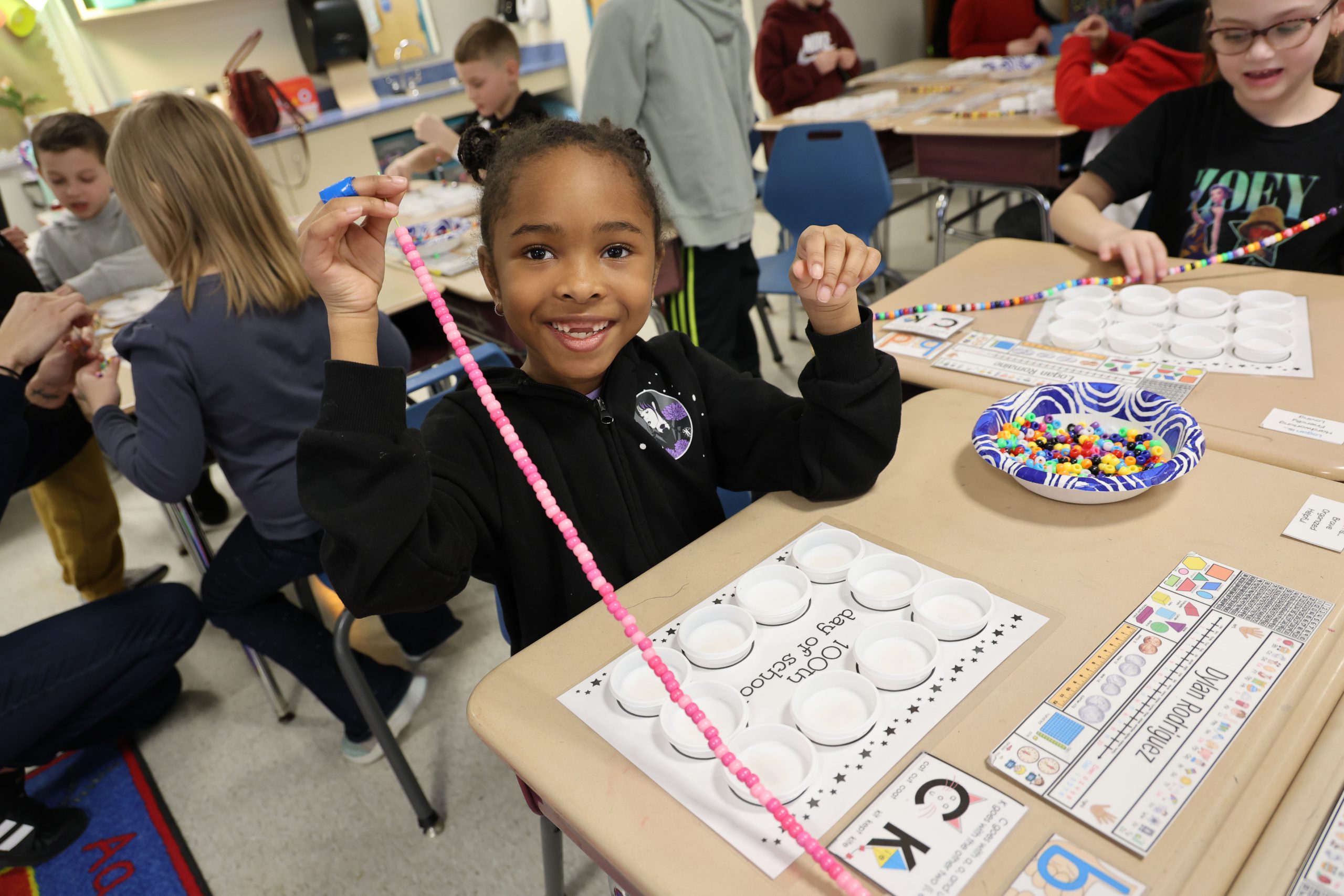 A young elementary girl smiles as she strings 100 pink beads.