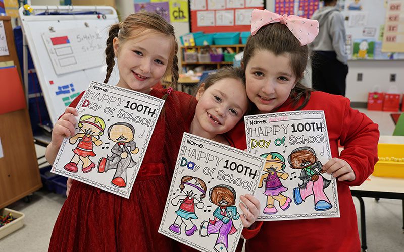 Three young elementary students hold up their coloring sheets celebrating 100 days of school.