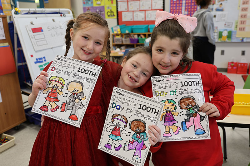 Three young elementary students hold up their coloring sheets celebrating 100 days of school.