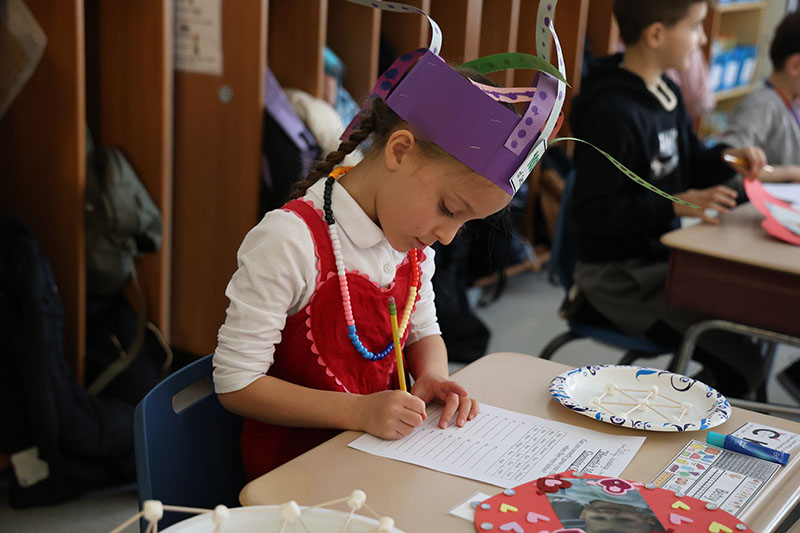 A second grade girl writes on a piece of paper as she looks at photos.