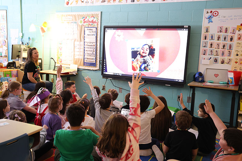 A class of second graders sit on a rug, many raising their hands to answer a question as a picture is displayed on the screen in front of them.