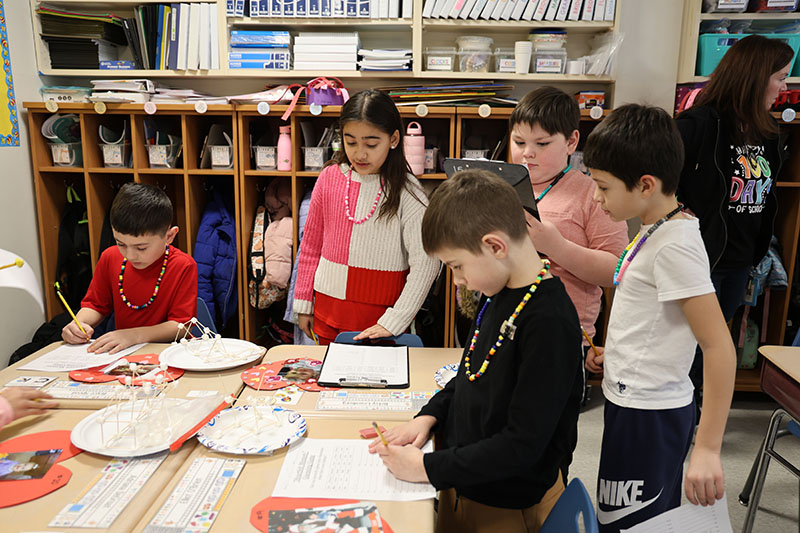 A group of second-grade boys and girls work around a bunch of desks.