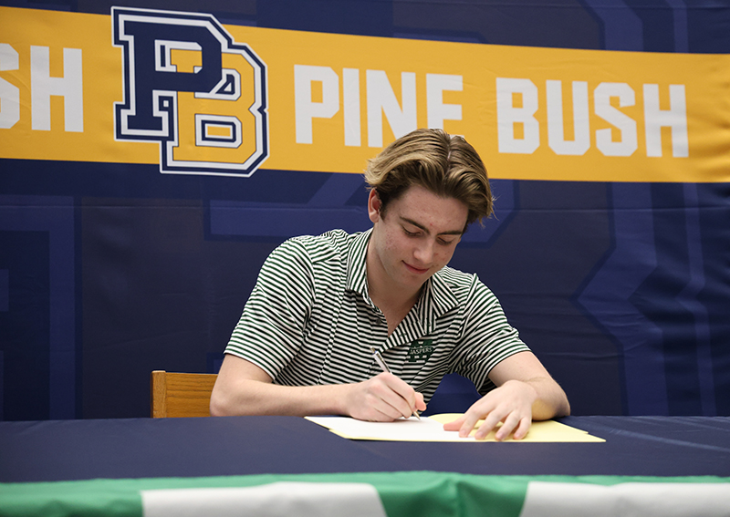 A high school senior sits at a table and signs a piece of paper. In the background is a blue and gold banner that says Pine Bush.