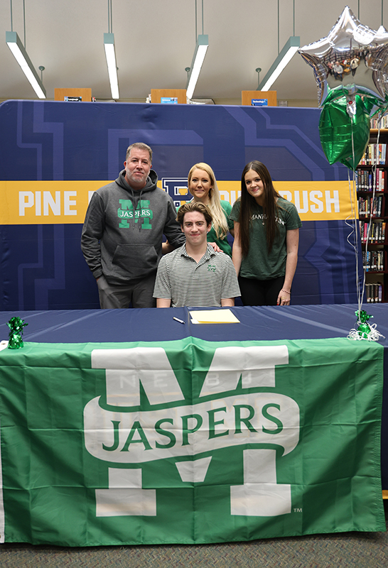 A high school senior sits at a table with a Jaspers banner on it. Behind him are a man and two women.