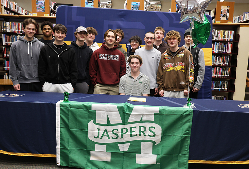 A high school senior sits at a table with a Jaspers banner on it. Behind him are 12 other high school boys.