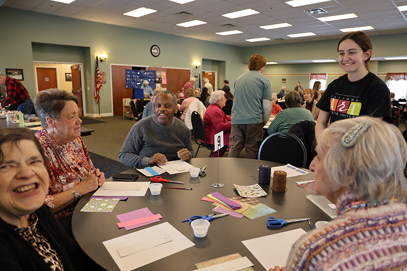 A high school girl stands at a table of senior citizens smiling and helping them do art projects.