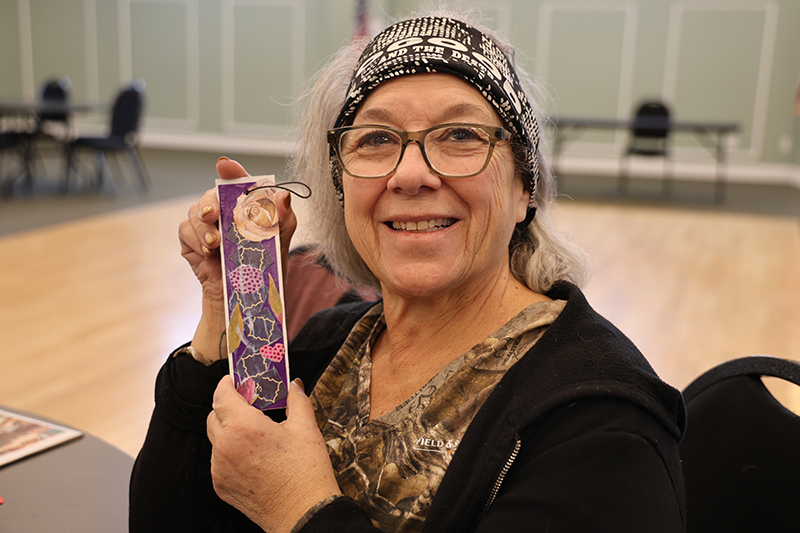 A woman holds up a bookmark she made. She is smiling.