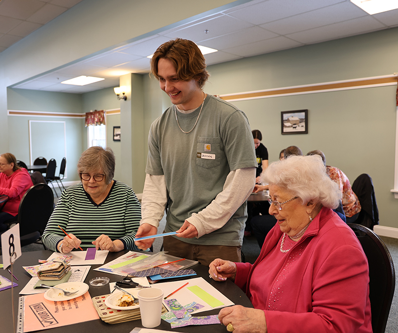 A high school boy smiles as he helps two women make art projects.