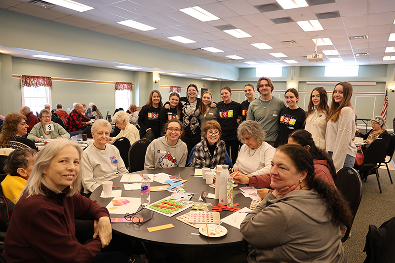 A group of 10 high school kids gather around a round table with senior citizens sitting at it. Everyone is smiling.