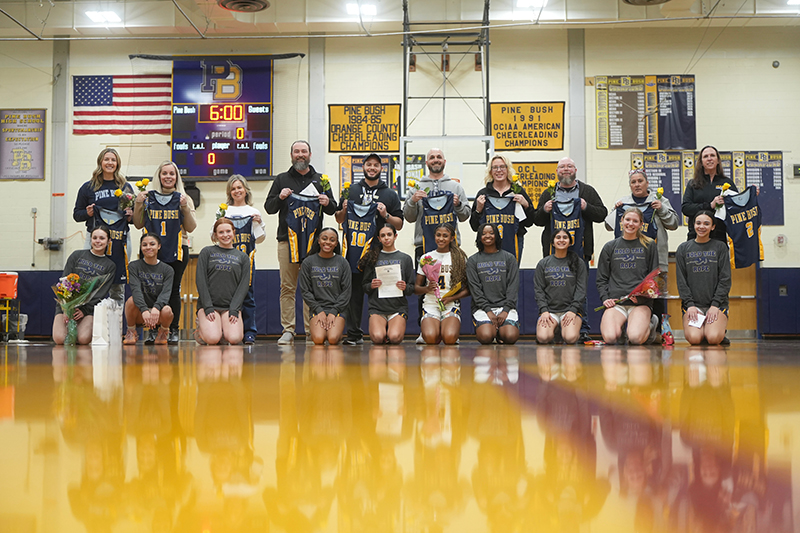 Ten high school students kneel in front and 10 adults stand behind them holding basketball jerseys and yellow roses. They are on a basketball court.