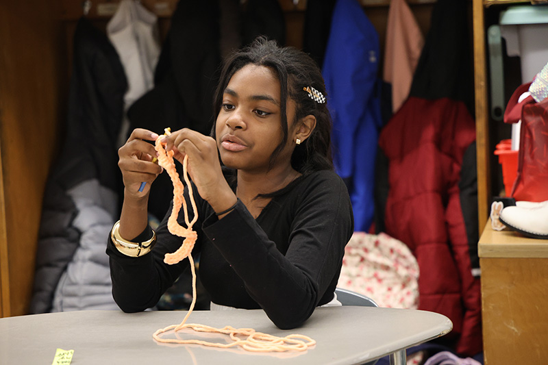 A girl with long dark hair sits at a desk and crochets.